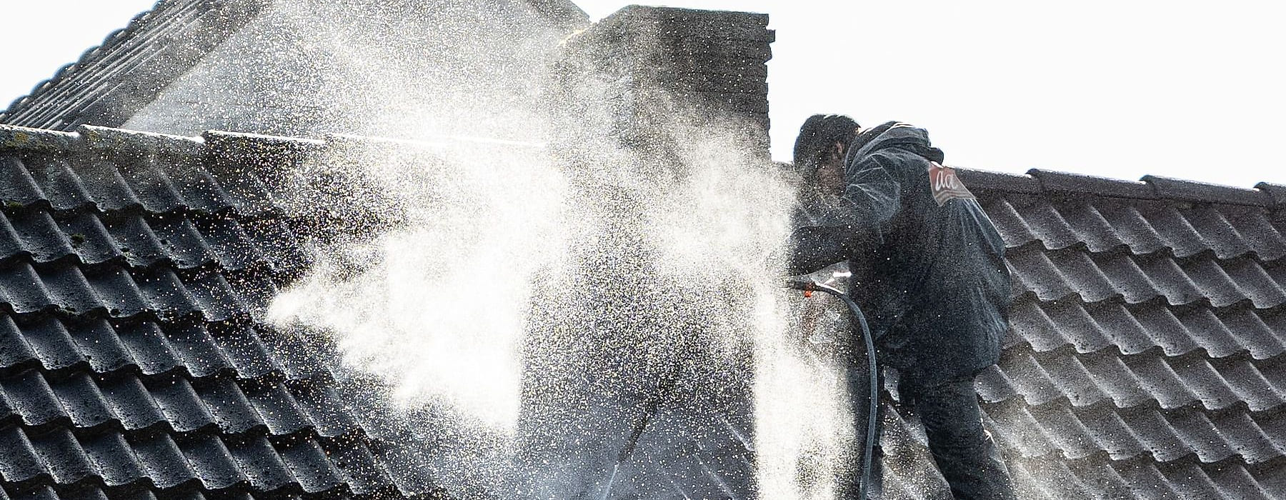 A technician on a tile roof washing it down, a plume of spray across the tiles